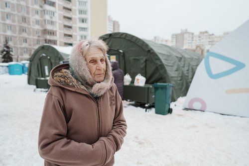 Vira Ivanivna, 75, steht vor dem Zelt einer staatlichen humanitären Hilfsstelle, während andere Einheimische sich dort aufwärmen und ihre Geräte aufladen. Dies geschieht während eines Stromausfalls, nachdem wichtige zivile Infrastruktur durch nächtliche russische Raketen- und Drohnenangriffe im Rahmen des russischen Angriffs auf die Ukraine getroffen wurde. Kyjiw, Ukraine, Februar 2026.