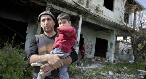 Ein Vater mit seinem Sohn im Dorf Khan Jouz. Im Hintergrund die ausgebrannte Ruine seines von der al-Nusra-Front zerstoertes Wohnhaus.