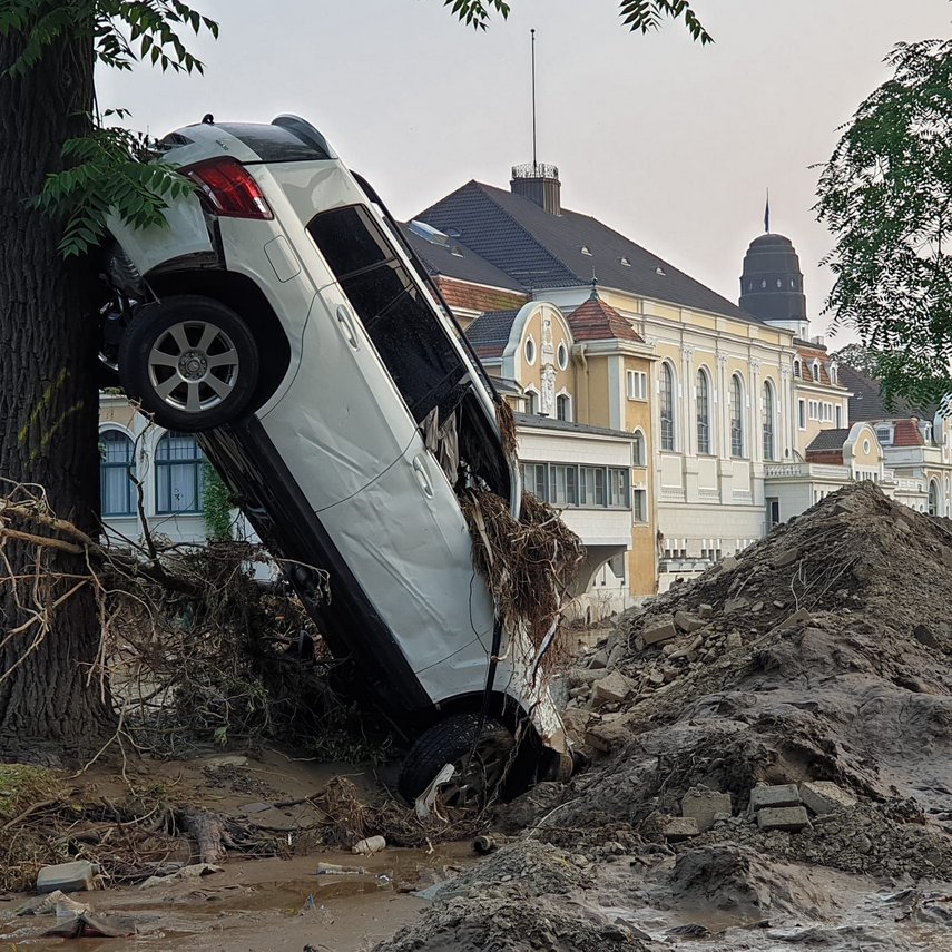 Ein demoliertes Auto steht hochkant an einem Baum, umgeben von Schlamm und Geröll. 